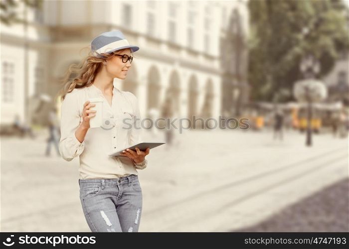 Beautiful young woman with a disposable coffee cup, drinking coffee, holding tablet in her hands, and crossing the street against urban city background.