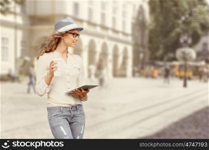 Beautiful young woman with a disposable coffee cup, drinking coffee, holding tablet in her hands, and crossing the street against urban city background.