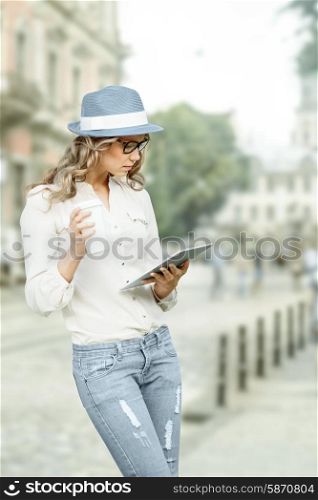 Beautiful young woman with a disposable coffee cup, holding tablet in her hands, reading news and studying against urban city background.