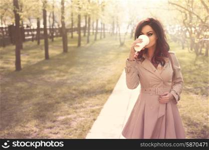 Beautiful young woman with a disposable takeaway cup and drinking coffee in a morning park, with the rising mist over trees and alleys.