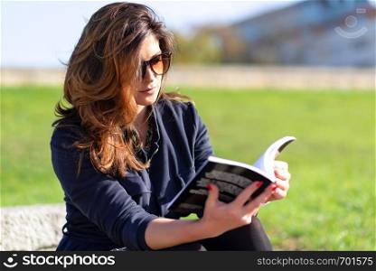 Beautiful young woman with a nice hairstyle and sunglasses sitting outdoor and reading a book on a sunny day.