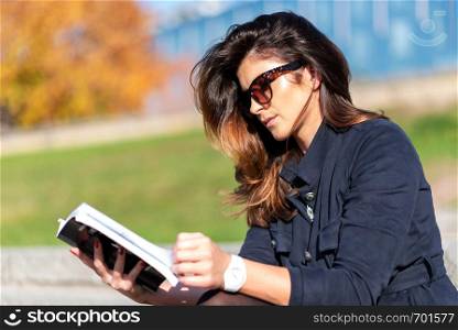Beautiful young woman with a nice hairstyle and sunglasses sitting outdoor and reading a book on a sunny day.