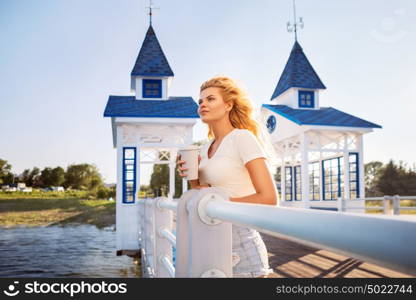 Beautiful young woman with a take away coffee cup, standing on the bridge near the water.