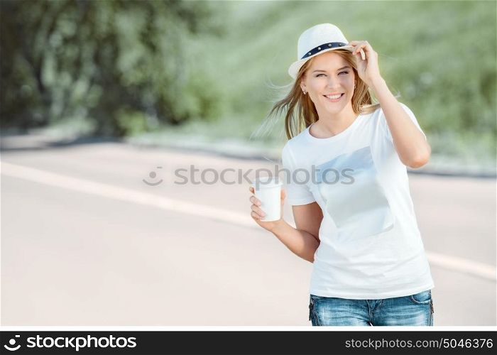 Beautiful young woman with a takeaway coffee cup, walking on the road, drinking coffee, and smiling against road background.