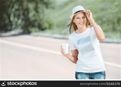 Beautiful young woman with a takeaway coffee cup, walking on the road, drinking coffee, and smiling against road background.