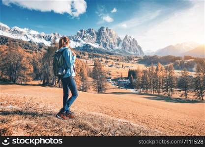 Beautiful young woman with backpack is standing on the hill against mountains at sunset in autumn. Landscape with sporty girl, rocks with snowy peaks, meadow, orange trees, blue sky in Italy. Travel