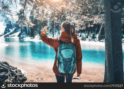 Beautiful young woman with backpack taking a selfie on the lake at sunset in autumn. Landscape with sporty girl is making photo, green trees, azure water in Dolomites, Italy. Travel in fall
