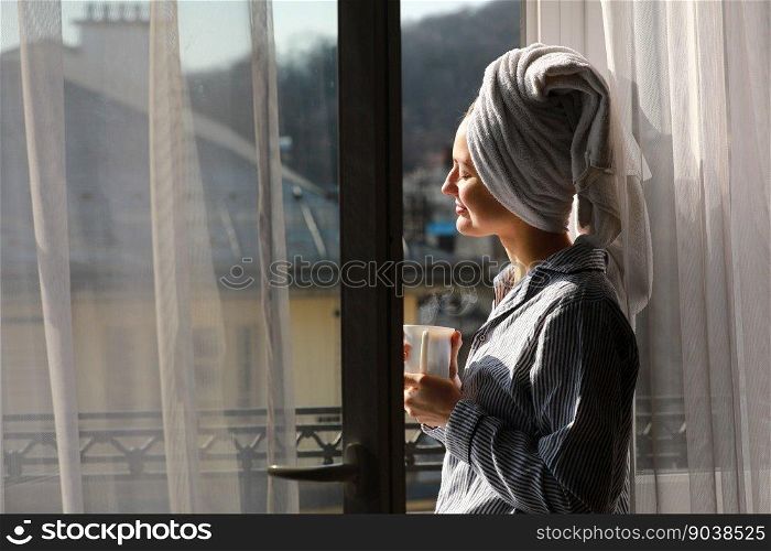 Beautiful young woman with bath towel on head holds a white cup of hot coffee or tea at home by the window. Smiling girl after shower is enjoying aroma drinks and have breakfast in the morning.. Beautiful young woman with bath towel on head holds a white cup of hot coffee or tea at home by the window. Smiling girl after shower is enjoying aroma drinks and have breakfast in the morning