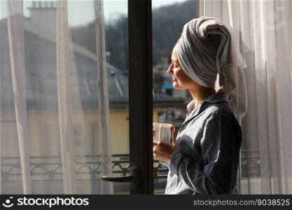 Beautiful young woman with bath towel on head holds a white cup of hot coffee or tea at home by the window. Smiling girl after shower is enjoying aroma drinks and have breakfast in the morning.. Beautiful young woman with bath towel on head holds a white cup of hot coffee or tea at home by the window. Smiling girl after shower is enjoying aroma drinks and have breakfast in the morning