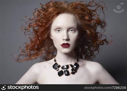 Beautiful young woman with black jewelry and red-hair on grey background. Nude pretty lady with bright red lipsstick and modern art makeup. Sensual model pose in photo-studio. Sexy naked body.