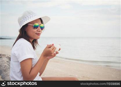 Beautiful young woman with coffee in hands sitting on the beach and watching the sunrise.