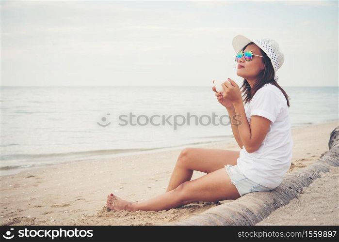 Beautiful young woman with coffee in hands sitting on the beach and watching the sunrise.
