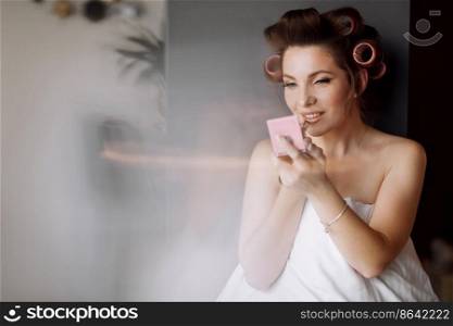 beautiful young woman with curlers is doing make-up while looking at the mirror on bed in the morning.. beautiful young woman with curlers is doing make-up while looking at the mirror on bed in the morning