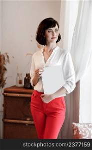 beautiful young woman with dark short hair holding a magazine with mock up. girl in a white shirt and red pants standing near the window.. beautiful young woman with dark short hair holding a magazine with mock up. girl in a white shirt and red pants standing near the window