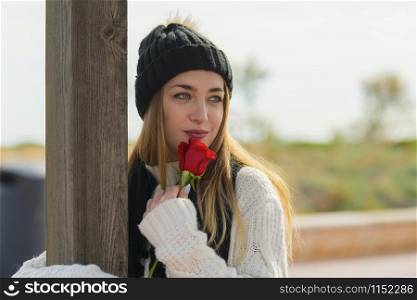 Beautiful young woman with dreamy eyes holds a red rose while looking away from the camera on an out of focus background. Valentine?s concept.. Beautiful young woman with dreamy eyes holding a rose