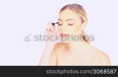 Beautiful young woman with her long blond hair in a ponytail and bare shoulders carefully applying mascara, isolated on white