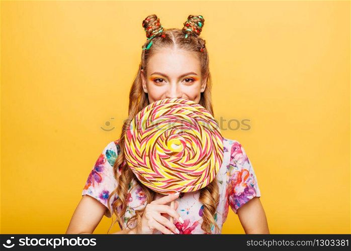 Beautiful young woman with huge candy instead of a head. Big colorful caramel lollypop in hands of woman. Portrait of attractive lady with big lollypop, yellow wall on background.. Woman with huge candy instead of a head