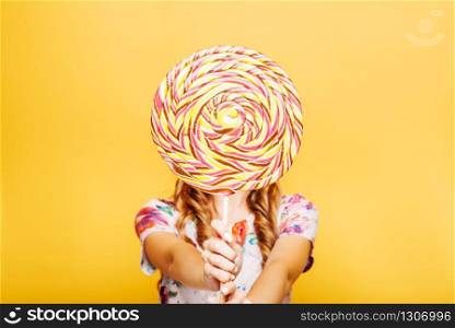 Beautiful young woman with huge candy instead of a head. Big colorful caramel lollypop in hands of woman. Portrait of attractive lady with big lollypop, yellow wall on background.. Woman with huge candy instead of a head