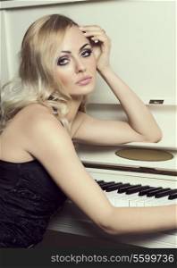Beautiful young woman , with long blond hair , in elegant black dress , near a piano looking in camera