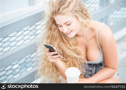 Beautiful young woman with long curly hair and a takeaway coffee cup, chatting and surfing on the phone against urban metal railing background.