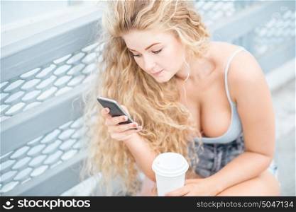 Beautiful young woman with long curly hair and a takeaway coffee cup, chatting and surfing on the phone against urban metal railing background.