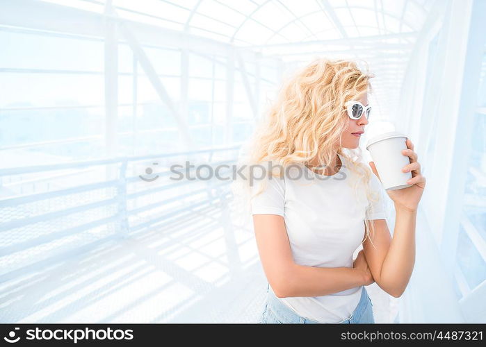 Beautiful young woman with long curly hair, holding a take away coffee cup and standing on the bridge against urban background.