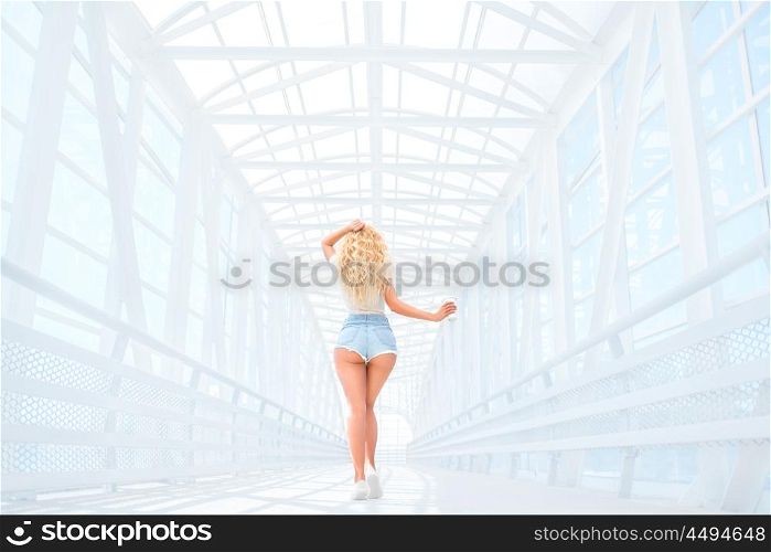 Beautiful young woman with long curly hair, holding a take away coffee cup and standing on the bridge against urban background, back view.