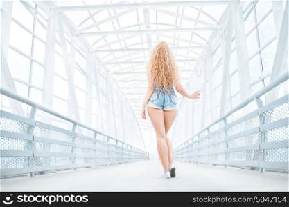 Beautiful young woman with long curly hair, holding a take away coffee cup and standing on the bridge against urban background, back view.