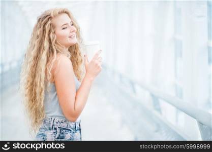 Beautiful young woman with long curly hair, holding a take away coffee cup and standing on the bridge against urban background.