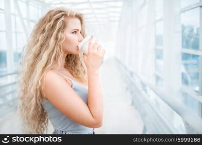 Beautiful young woman with long curly hair, holding a take away coffee cup and standing on the bridge against urban background.