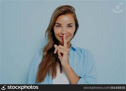 Beautiful young woman with long healthy hair showing shush gesture while mysteriously smiling at camera, asking to keep gossip in secret on light blue background. Body language concept. Beautiful woman showing shush gesture on light blue background