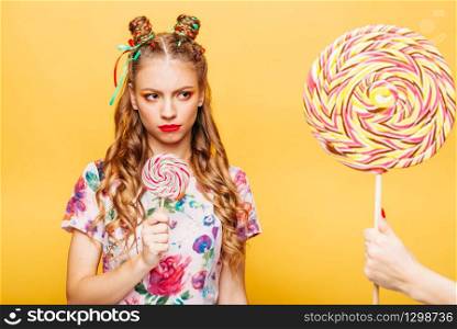 Beautiful young woman with mistrustful look holds in hand big lollypop. Somebody gives one more candy to girl, studio portrait. Stylish girl in summer colorful dress, yellow wall on background.. Stylish girl in summer colorful dress