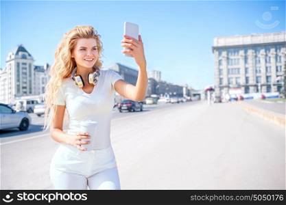 Beautiful young woman with music headphones and a take away coffee cup, taking picture of herself, selfie against urban city background.