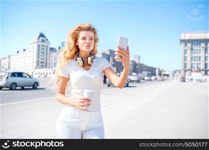 Beautiful young woman with music headphones and a take away coffee cup, taking selfie and making duck face against urban city background.