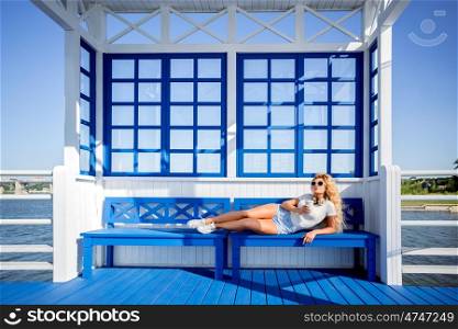 Beautiful young woman with music headphones and take away coffee cup, lying on the bench and waiting for a water taxi.
