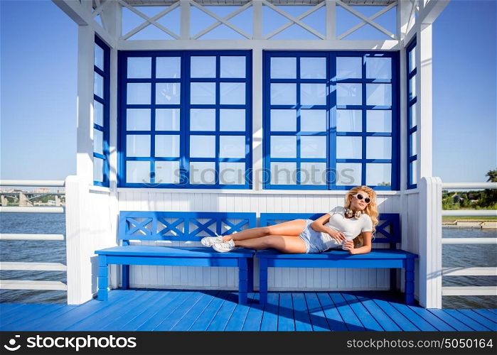 Beautiful young woman with music headphones and take away coffee cup, lying on the bench and waiting for a water taxi.