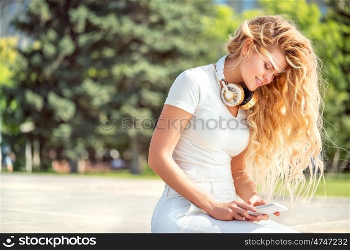 Beautiful young woman with music headphones around her neck, surfing internet on a smartphone and sitting against park background.