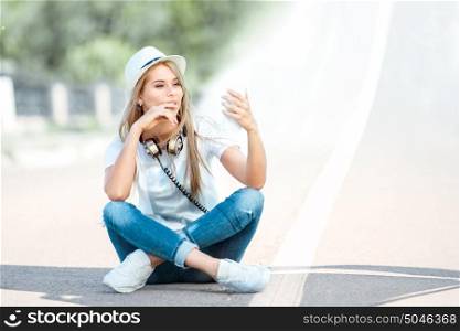 Beautiful young woman with music headphones around her neck, surfing internet on a smartphone and sitting on a separating strip on the road.