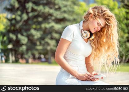 Beautiful young woman with music headphones around her neck, surfing internet on a smartphone and sitting against park background.