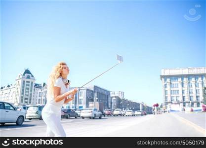 Beautiful young woman with music headphones around her neck, taking picture of herself with selfie stick against urban city.