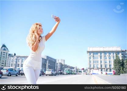 Beautiful young woman with music headphones around her neck, taking picture of herself, selfie against urban city background.