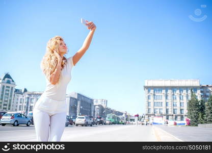 Beautiful young woman with music headphones around her neck, taking picture of herself, selfie against urban city background.