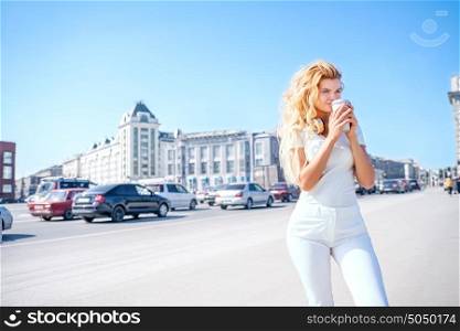Beautiful young woman with music headphones drinking from a take away coffee cup and posing against urban city background.