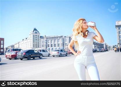 Beautiful young woman with music headphones drinking from a take away coffee cup and posing against urban city background.