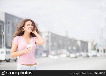 Beautiful young woman with music headphones, holding a take away coffee cup, listening to the music and looking aside against city traffic background.