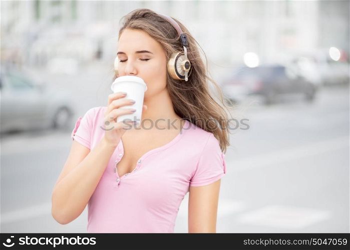 Beautiful young woman with music headphones, holding a take away coffee cup and listening to the music with her eyes closed against city traffic background.