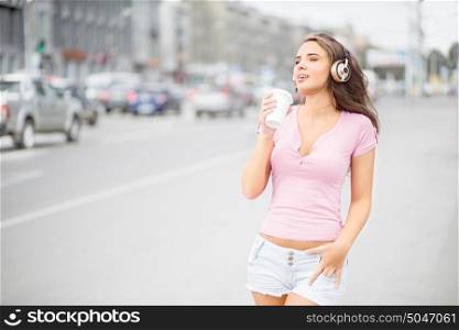 Beautiful young woman with music headphones, holding a take away coffee cup and listening to the music with her eyes closed against city traffic background.