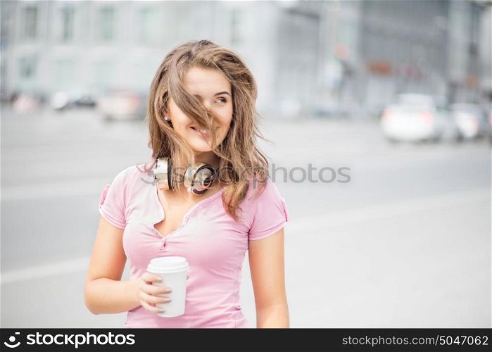 Beautiful young woman with music headphones, holding a take away coffee cup, listening to the music and looking aside against city traffic background.