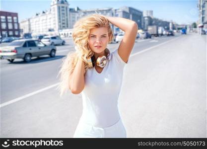 Beautiful young woman with music headphones on her neck, standing against urban city background and looking straight to the camera.