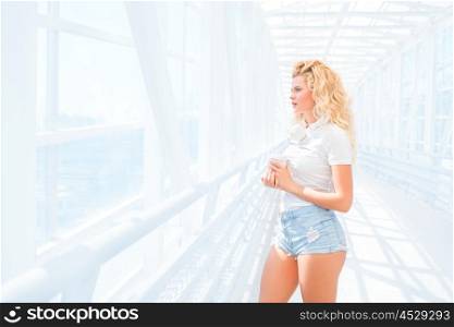 Beautiful young woman with music headphones, standing on the bridge with a take away coffee cup and posing against urban background.